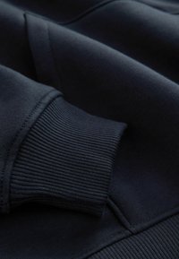 Mørkeblå sweatshirt-stof med ribbede manchetter og en glat tekstur. Har kontrasterende syninger langs sømmene for øget holdbarhed.