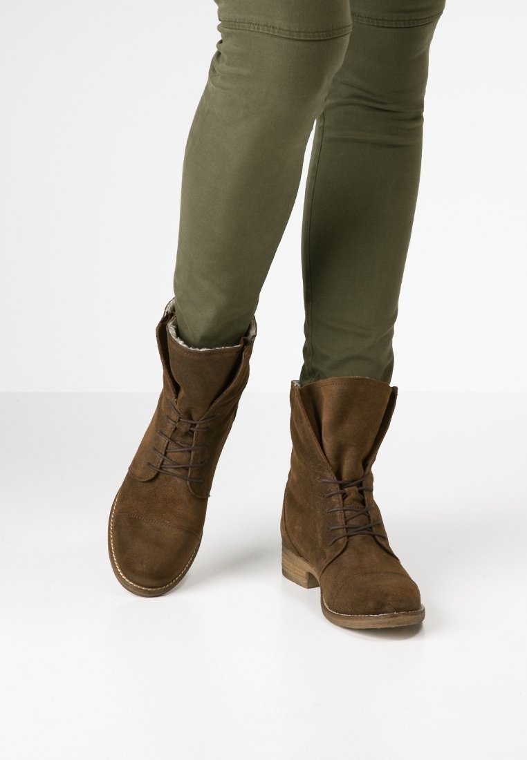 Bottes basses en daim marron au design décontracté, fermeture à lacets, et petit talon en bois, portées avec un pantalon ajusté vert olive.
