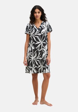 Vrouw, blootsvoets, gekleed in een zwart jurkje met korte mouwen, grote witte bloemenprint en een V-hals, staand tegen een effen witte achtergrond.