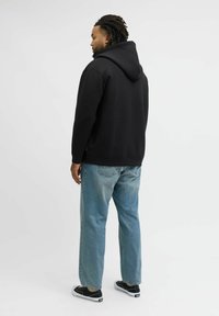 Felpa nera con cappuccio e tasca frontale, abbinata a jeans azzurri dal taglio morbido e sneakers nere, vista da dietro.