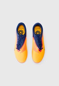 Chaussures de soccer orange vif et marine avec un dessus texturé, un motif géométrique et des lacets, présentant une semelle intérieure jaune avec un logo.