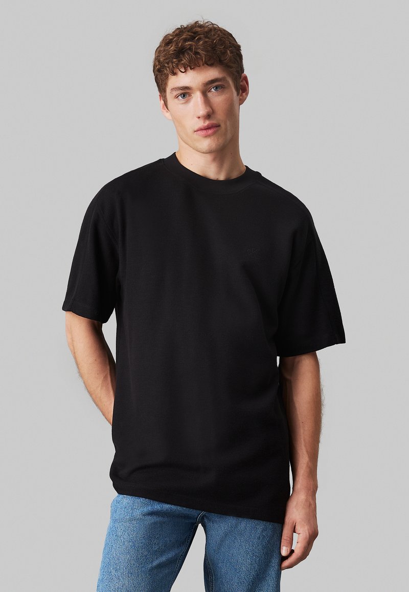 T-shirt nera a maniche corte con scollatura rotonda, realizzata in un tessuto texturizzato. Presenta un piccolo logo sul lato sinistro del petto. Indossata con jeans blu.