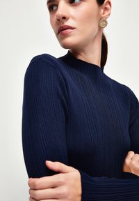 Marinblå ribbade tröja med hög hals och långa ärmar. Har en texturerad tyg och en kroppsnära passform, prydd med ett runt guldörhänge.