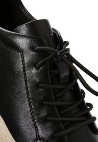 Zbliżenie na czarny skórzany sneaker z okrągłymi czarnymi sznurowadłami zabezpieczonymi czarnym blokadą sznurowadeł oraz beżową gumową podeszwą widoczną na krawędzi.