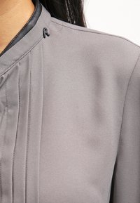 Grå bluse med en glat tekstur, plisseret design og en sort læderkrave med en lille sort detalje nær udskæringen.