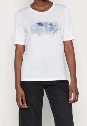 Osoba ubrana w białą koszulkę z krótkim rękawem z niebieskim pociągnięciem pędzla i słowem „enjoy” zapisanym kursywą, w połączeniu z czarnymi spodniami.