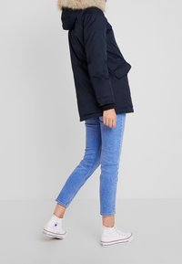 Marinblå puffjacka med pälsbesatt huva, sidofickor och ribbade ärmmuddar, som bärs med ljusblå jeans och vita högsta sneakers.