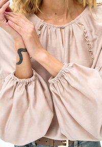 Kobieta w beżowej bluzce z falbankami na rękawach dotyka nadgarstka z tatuażem czarnej falistej linii, nosi brązowy pasek i niebieskie dżinsy.