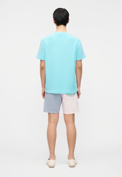 Άνδρας που στέκεται με την πλάτη, φοράει ανοιχτό μπλε κοντομάνικο πουκάμισο, πολύχρωμο ριγέ σορτς και λευκά παπούτσια χωρίς κορδόνια.