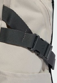 Mochila de tela beige con correas de cincha gris oscuro y una hebilla de plástico negra con un logotipo. Incluye detalles de costuras reforzadas.