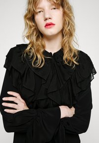 Blusa nera con strati di volant sul colletto, maniche lunghe con polsini arricciati e una texture di tessuto liscia e morbida.