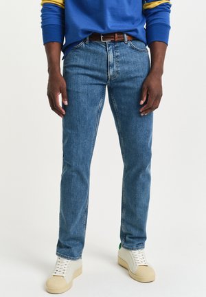 Mann som har på seg blå jeans, brun belte, hvite joggesko og en blå hettegenser med gule striper på ermene, stående på en hvit bakgrunn.