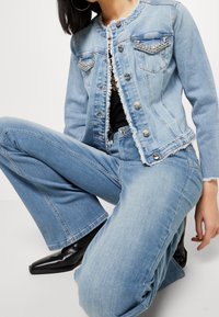 Mujer de rodillas vistiendo una chaqueta de denim de color azul claro con tachuelas plateadas y unos pantalones de mezclilla acampanados de cintura alta a juego, junto con botas negras de punta.
