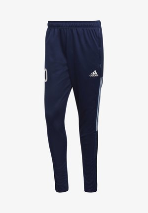 Mann trägt dunkelblaue Adidas-Trainingshose mit der Nummer 10 am linken Oberschenkel und schwarze Sportschuhe, steht mit einer Hand in der Tasche.