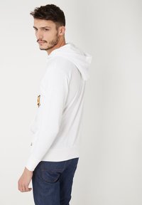 Sudadera blanca con bolsillo frontal, puños y dobladillo acanalados. Presenta un emblema gráfico en la manga izquierda. Combinada con jeans de mezclilla oscura.
