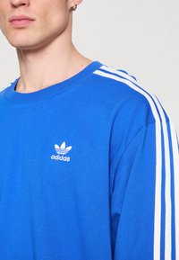 Kék pamut pulóver fehér Adidas logóval és három fehér csíkkal a vállakon, kerek nyakkivágással és puha textúrával.