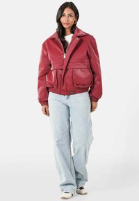 Rode faux leren jas met een brede kraag, elastische zoom en grote zakken aan de voorkant, gecombineerd met lichtgewassen, wijde jeans en sneakers.