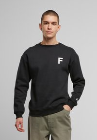 Must valguskate, millel on ümar kaelus, millel on rinnas valge "F" logo ja tikand varruka otstes. Mugav kangas.