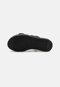 Semelle de chaussure en caoutchouc noir avec une surface texturée et des rainures horizontales. Présente un petit logo sur le côté pour l'identification de la marque.