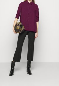 Camicia viola a maniche lunghe con bottoni davanti, pantaloni neri svasati e stivaletti neri con tacco. Tiene in mano una borsa rotonda a fantasia scozzese.
