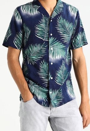 Mężczyzna ubrany w granatową koszulę z krótkim rękawem zapinaną na guziki z zielonym nadrukiem liści palmowych oraz jasnoszare spodnie, ręce częściowo w kieszeniach.