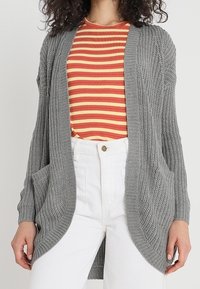 Mujer con un cárdigan gris de punto sobre una camisa a rayas roja y amarilla, con pantalones blancos de cintura alta, con las manos relajadas a los lados.