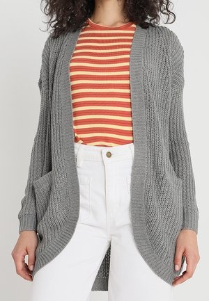Femme portant un cardigan en tricot gris sur une chemise à rayures rouges et jaunes avec un pantalon taille haute blanc, les mains détendues le long du corps.