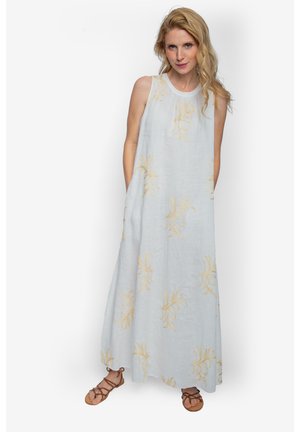 Kobieta z falującymi blond włosami, ubrana w długą, białą sukienkę bez rękawów z żółtymi kwiatowymi wzorami oraz brązowe, płaskie sandały na paski.