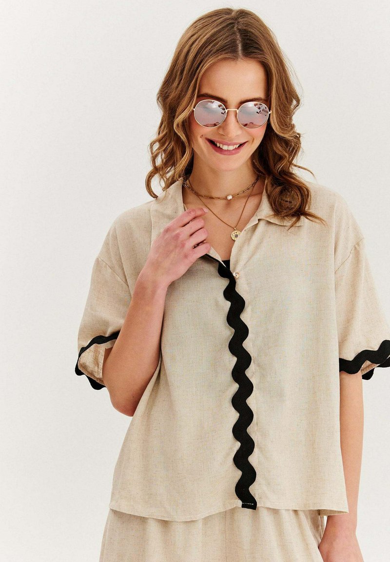Béžová košile na knoflíky s černým vlnkovaným lemem, krátké rukávy, volný střih a texturovaná tkanina; doplněná kulatými růžovými slunečními brýlemi.