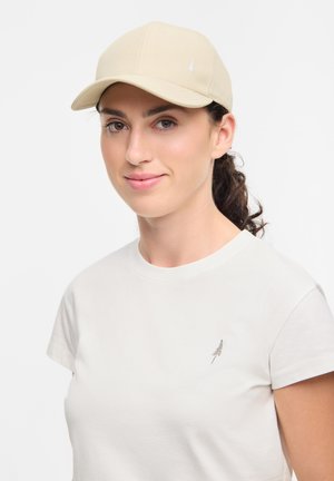Beige pet met een gebogen rand, gemaakt van zachte stof, met een subtiele boomlogo geborduurd op de voorkant. Gekocht met een eenvoudig wit T-shirt.