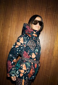 Kolorowa, wzorzysta kurtka puchowa z motywami kwiatowymi, geometrycznymi i zwierzęcymi. Wysoki kołnierz, pasek w talii, noszona z dużymi okularami przeciwsłonecznymi.