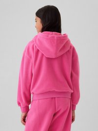 Tânărul cu păr lung și închis la culoare poartă un hanorac roz deschis din fleece și pantaloni asortați, întors cu spatele, pe un fundal gri simplu.