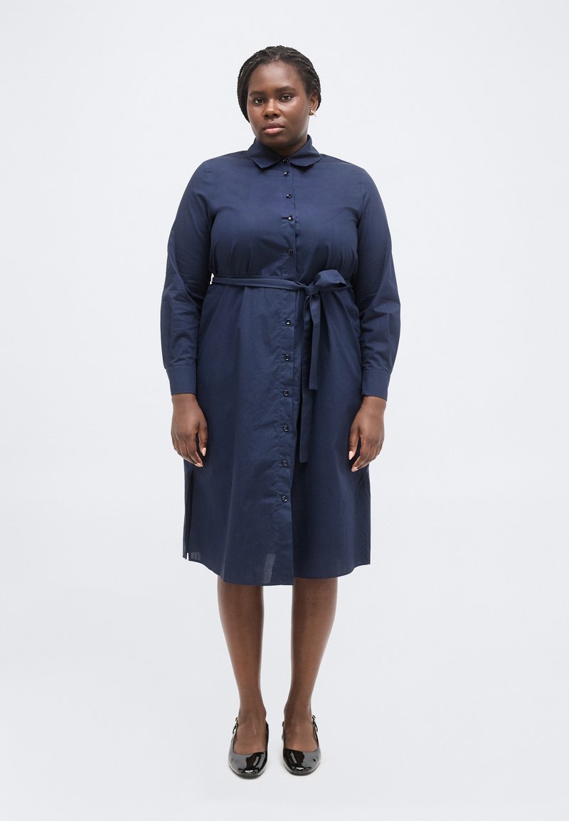 Γυναίκα που στέκεται φορώντας ένα μπλε ναυτικό πουκάμισο-φόρεμα με κουμπιά και ζώνη στη μέση, και μαύρα παπούτσια από πατινέ δέρμα σε λευκό φόντο.