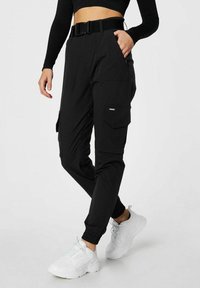 Person iført sorte cargobukser med elastiske manchetter, sort bælte, sort langærmet croppet top og hvide chunky sneakers, stående med den ene hånd i lommen.