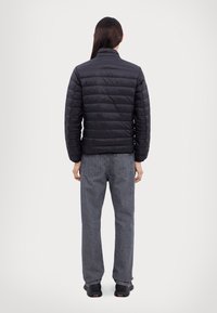 Personne aux longs cheveux foncés portant une veste matelassée noire, un jean gris et des chaussures noires, debout de dos sur un fond uni.
