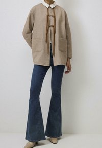 Jasnobrązowa kurtka z mieszanki wełny z dużymi przednimi kieszeniami, zawiązywana na plecione sznurki. Nosi się ją na białej koszuli z kołnierzykiem i w połączeniu z rozszerzającymi się ciemnoniebieskimi dżinsami.