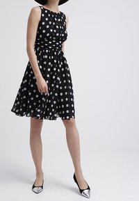 Robe noire à pois avec un corsage ajusté et une jupe évasée, en tissu léger. Le mannequin porte des talons noirs avec des accents argentés.