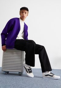 Cardigan tricotat mov cu nasturi negri, tricou alb, pantaloni largi negri și adidași albi cu accente negre pe o valiză argintie.