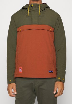 Mann trägt zweifarbige Kapuzenjacke mit olivgrünen Ärmeln und oberer Brust, rostorangefarbenem Frontpanel, gelben Druckknöpfen und Kordeln.