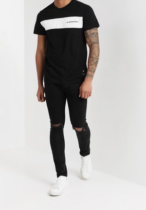 Schwarzes T-Shirt mit weißem horizontalem Streifen und "G-STAR RAW"-Logo. Kombiniert mit zerrissenen schwarzen Jeans und weißen Turnschuhen.