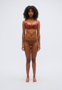 Soutien-gorge rouge et imprimé léopard avec bonnets rembourrés, bretelles réglables, et un bas de bikini marron assorti. Le mannequin est pieds nus sur un fond neutre.
