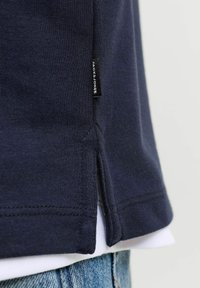 Gros plan sur l'ourlet en tissu bleu marine avec une fente latérale et une étiquette noire sur un vêtement bleu foncé porté sur une chemise blanche et un jean bleu.