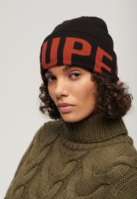 Vrouw met krullend haar, die een zwarte beanie met rode letters en een olijfgroene kabelgebreide coltrui draagt, tegen een effen achtergrond.