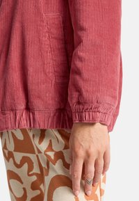 Giacca di velluto a coste rosa con una superficie testurizzata, polsini elasticizzati e una tasca laterale, abbinata a pantaloni beige e marroni con motivo.