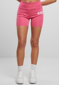 Rozā sporta šorti ar gludu tekstūru un augstu jostasvietu. Sānos ir balts uzraksts "KAN". Valkāti ar baltiem sporta apaviem un zeķēm.