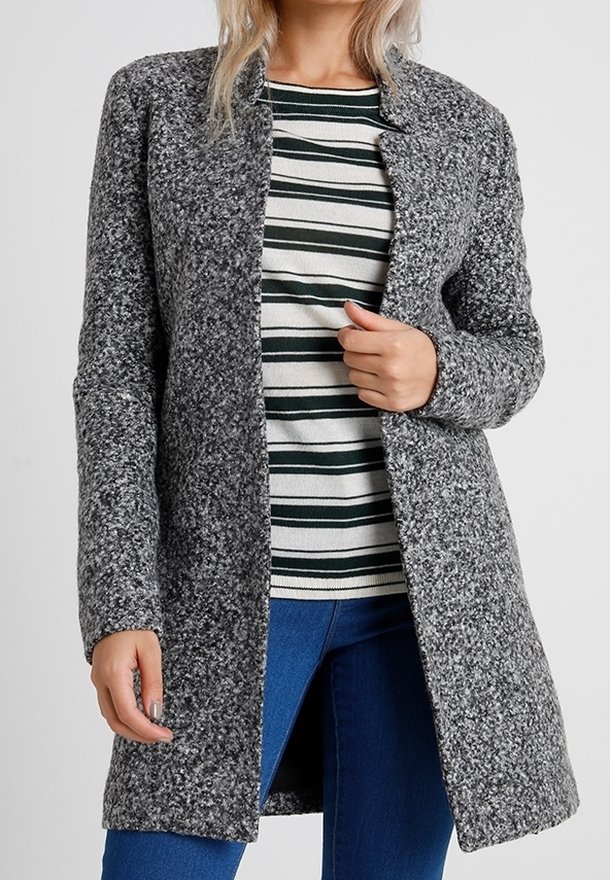 Femme portant un manteau gris texturé ouvert sur le devant, par-dessus une chemise rayée blanche et vert foncé, et un jean bleu, sur un fond uni.