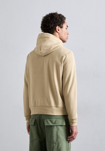 Haut à capuche beige avec cordon de serrage, poignets et ourlet côtelés, associé à un pantalon cargo vert avec poches latérales et un détail de bouton.