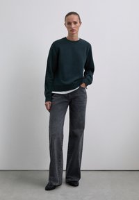 Mujer de pie contra un fondo liso, vistiendo un sudadera verde oscuro, una camisa clara debajo, pantalones anchos gris y zapatos negros.