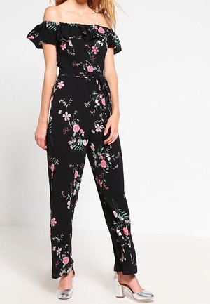 Jumpsuit noir à fleurs avec encolure smockée dégageant les épaules, présentant des fleurs roses et des feuilles vertes, associé à des chaussures argentées à talons carrés brillants.
