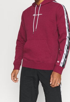 Mann trägt einen dunkelroten Champion-Hoodie mit weißem Logoschriftzug und weißen Logobändern entlang der Ärmel sowie schwarze Hose.